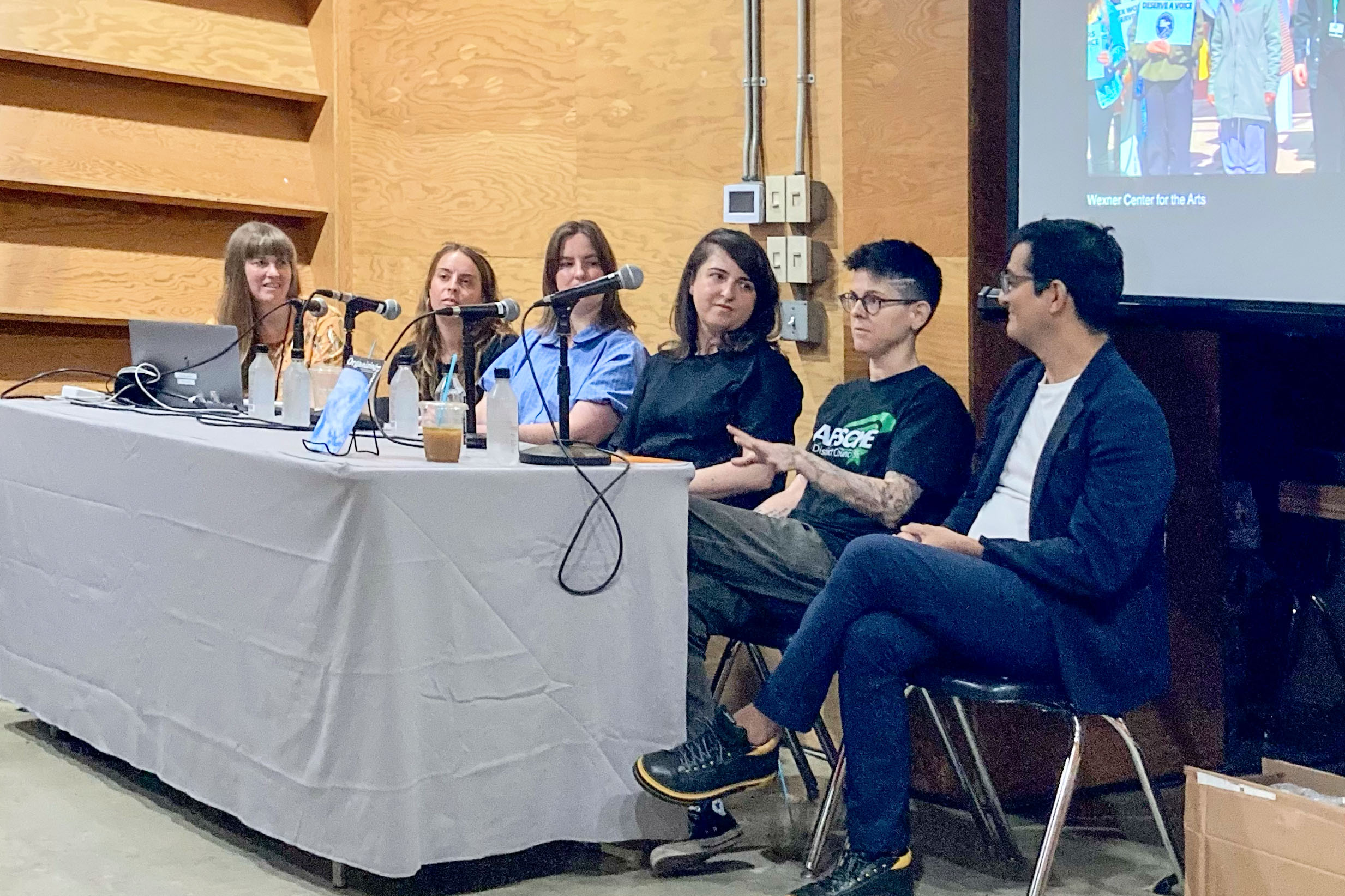 Six panelists seated at a table with microphones during the Organizing Power panel discussion at the Los Angeles Art Book Fair, August 2023. A projected slideshow showing union organizing imagery is visible in the background.