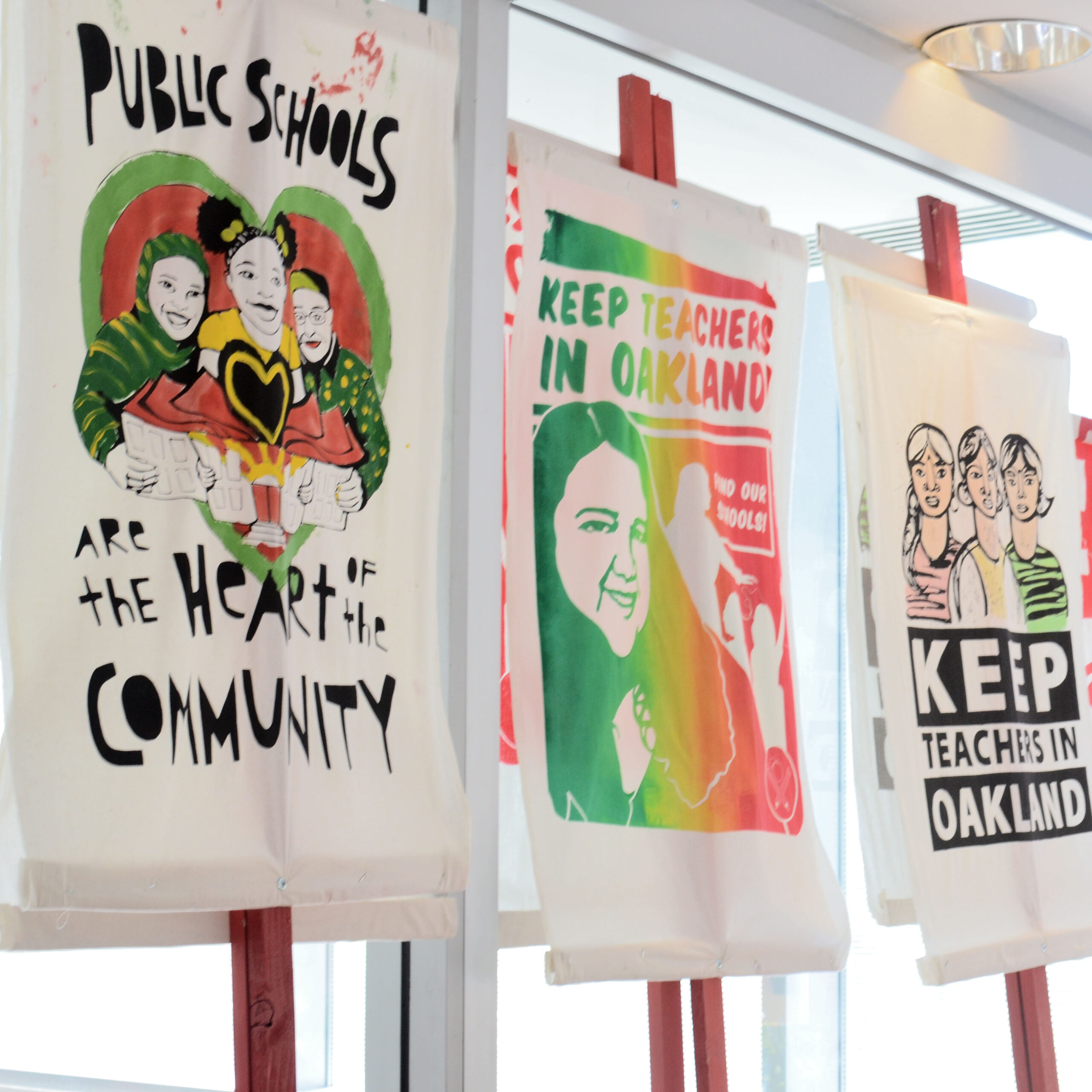 Colorful hand-painted banners hanging on wooden easels at the 2019 YBCA Public Square event. The banners read 'Public Schools are the Heart of the Community' and 'Keep Teachers in Oakland,' created during the 2019 Oakland teachers' strike.