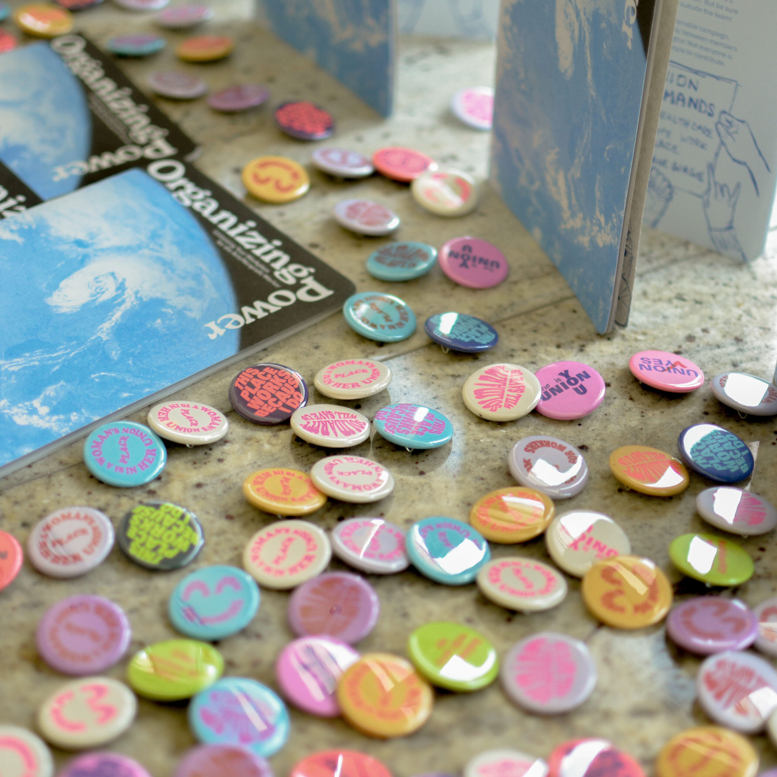 A scattered collection of colorful pro-union pinback buttons in pink, teal, green, and gold, displayed alongside copies of the Organizing Power booklets on a table at the 2019 YBCA Public Square event. Button slogans include 'Union Yes' and 'This Place Needs a Union.'