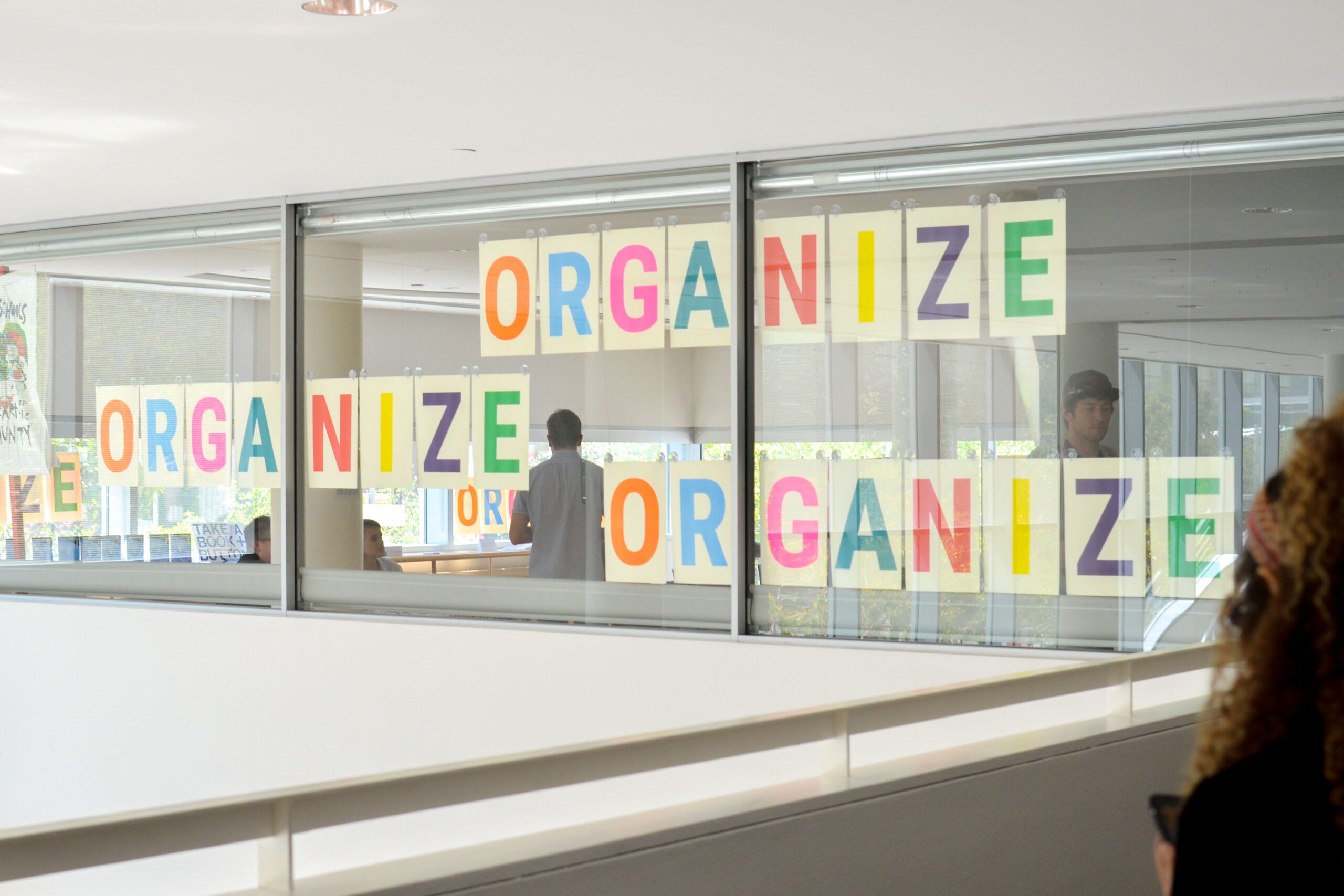 Colorful "ORGANIZE" signs displayed in the windows of the conference room, viewed from an interior hallway with people visible inside.