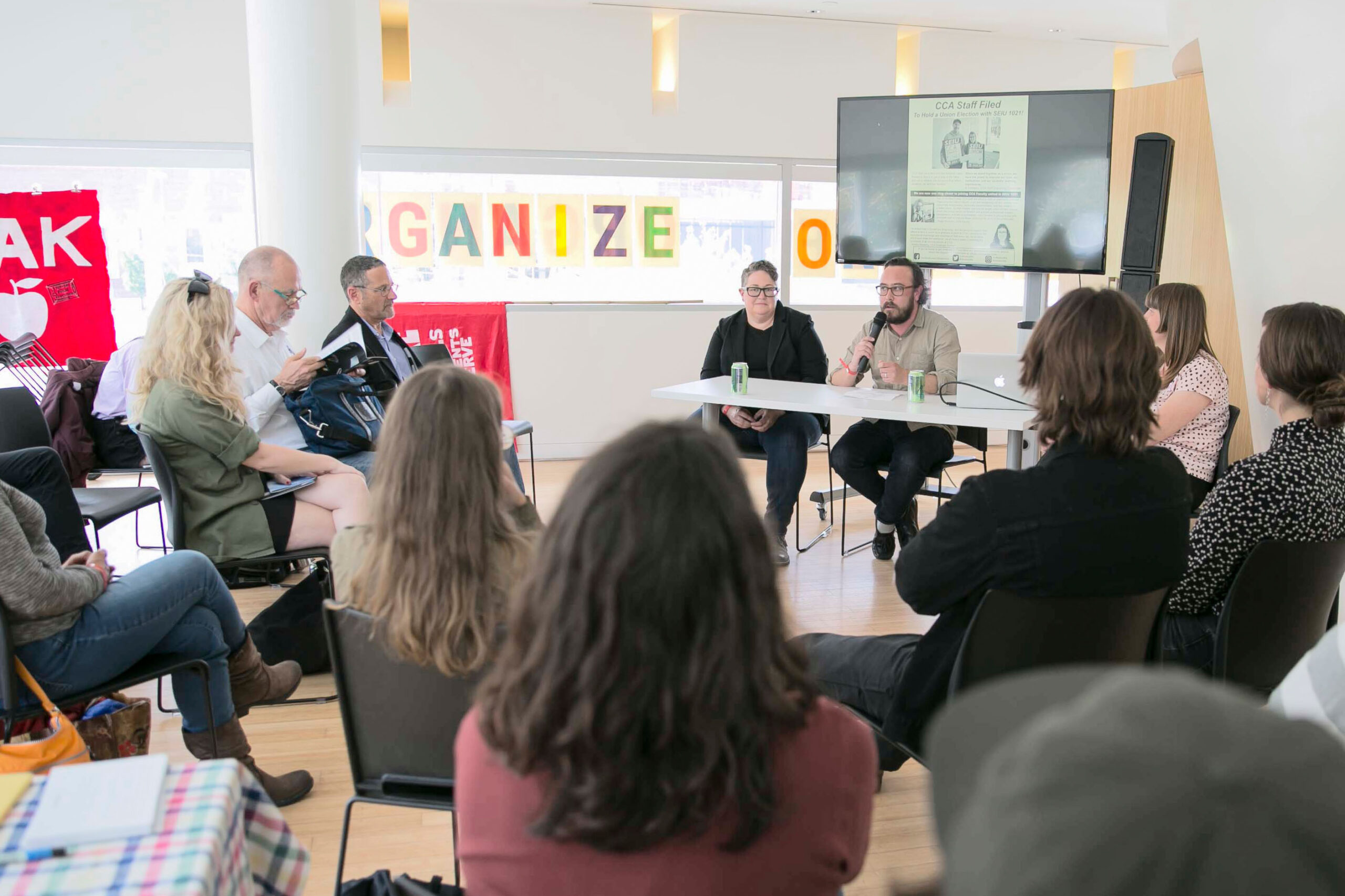 Attendees seated in a circle during a roundtable discussion at the Organizing Power Public Square event at YBCA, June 2019. Two speakers sit at a table at the front of the room holding a microphone, with the colorful 'ORGANIZE' window installation and Oakland teachers' strike banners visible in the background. A screen displays a CCA union organizing flyer.